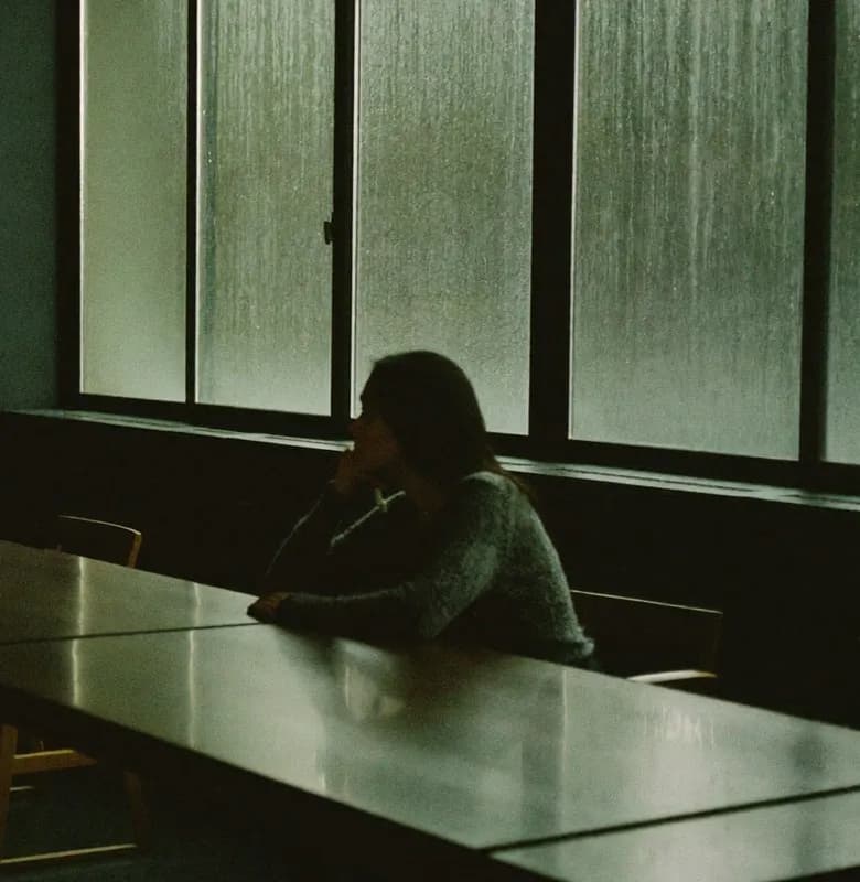 A person sitting alone at a long table beside a tall window in a dim room