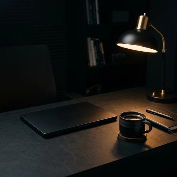 A lamp-lit desk with a closed laptop and a cup of coffee