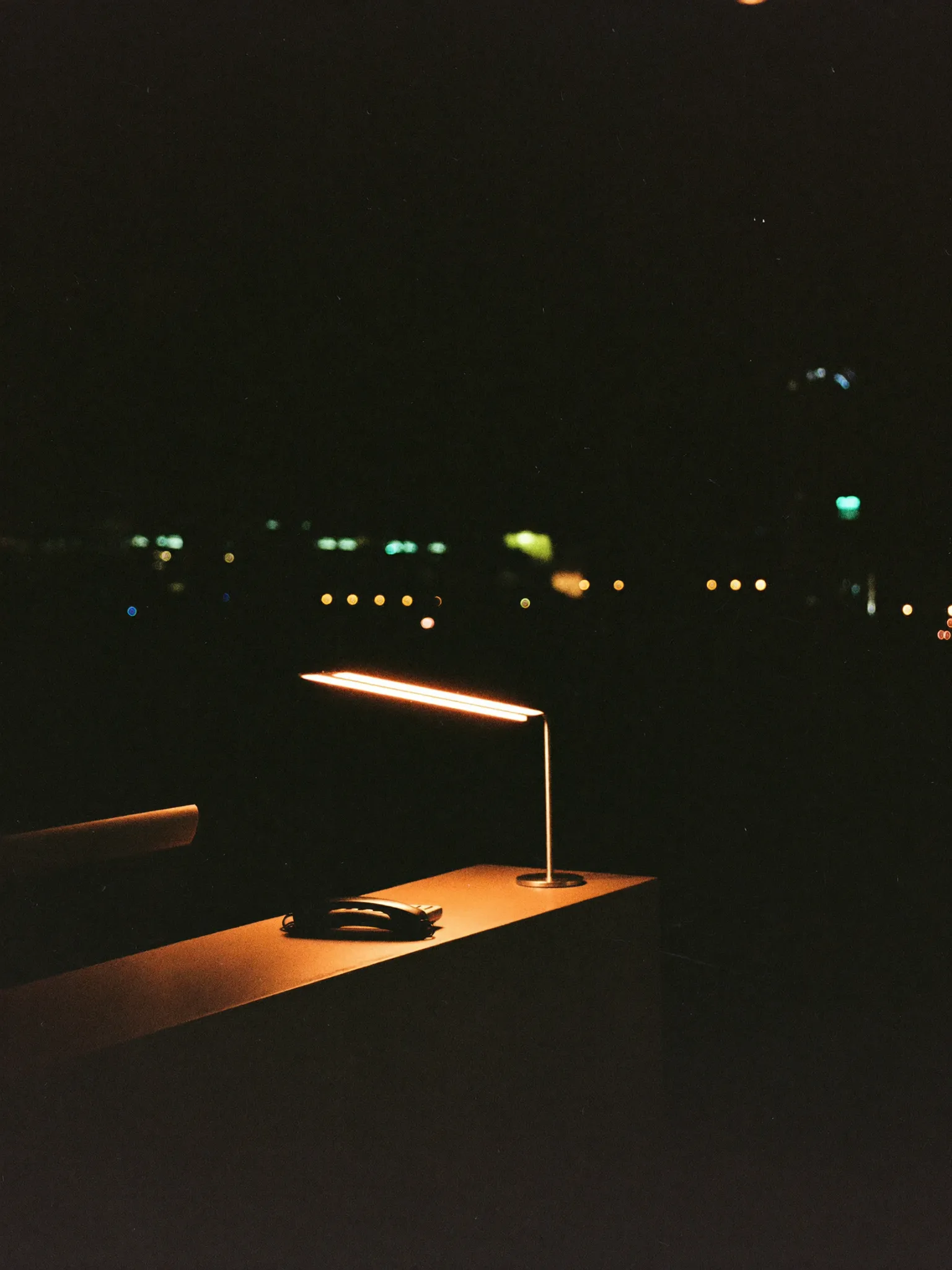 An empty reception desk late at night, lit by a single lamp, a telephone on the counter and city lights beyond the window