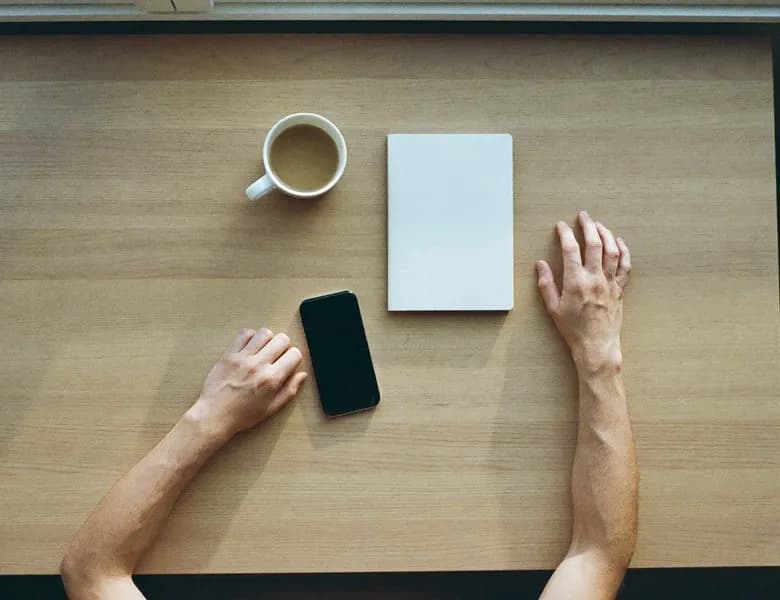 Overhead view of hands on a wooden desk beside a phone, a notebook and a coffee cup