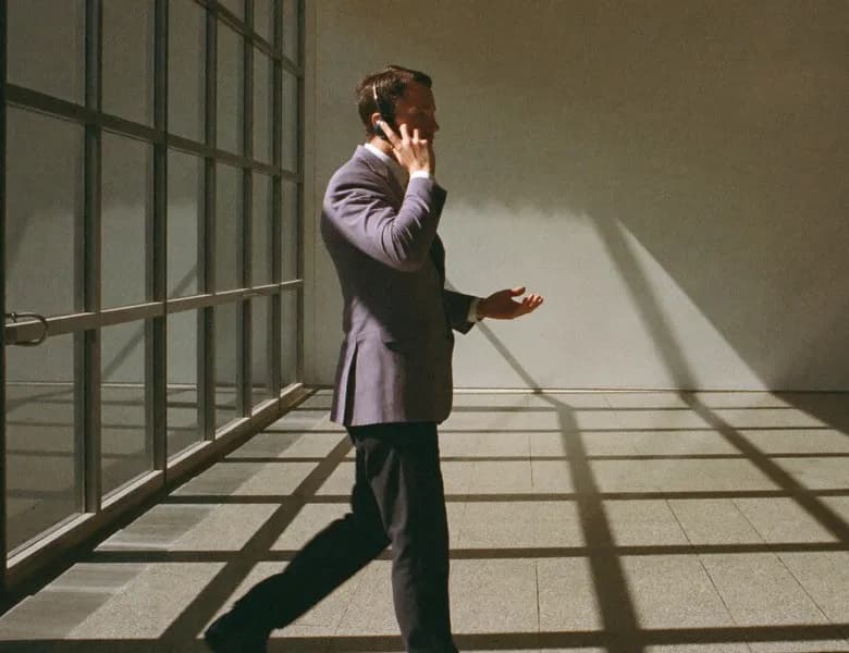 A man in a suit talking on the phone in a sunlit room with tall windows