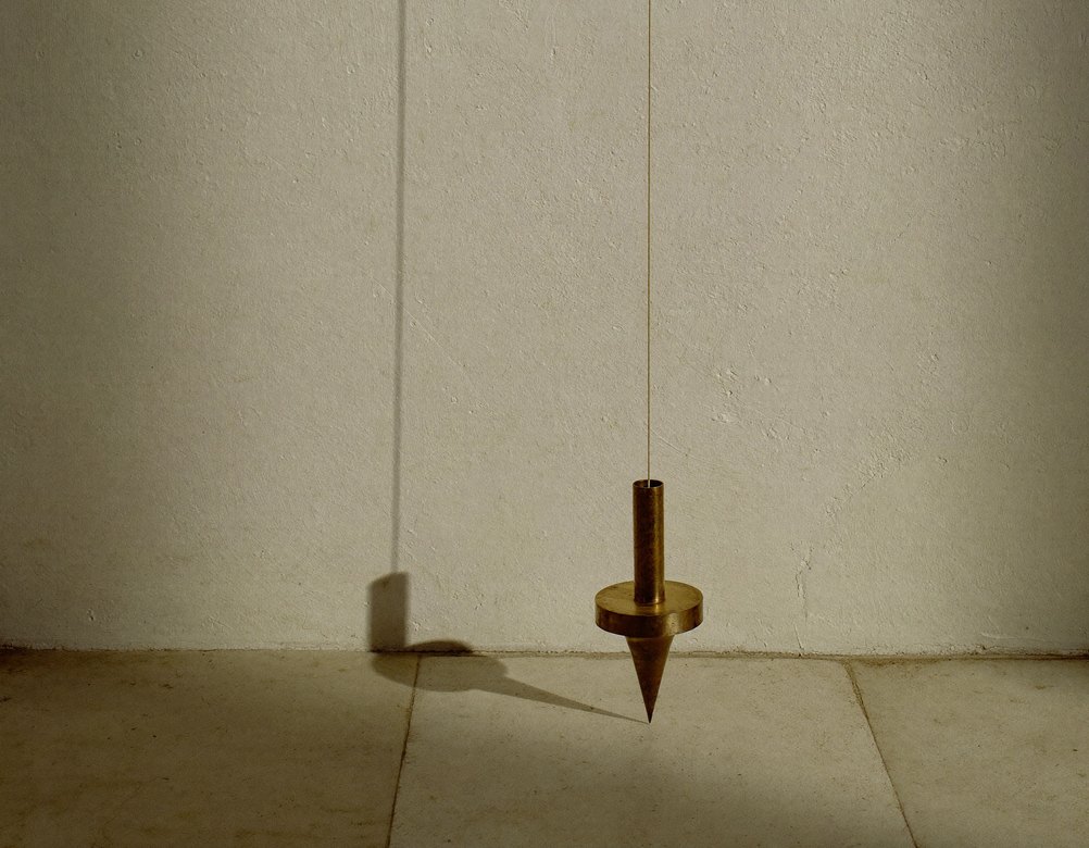A brass plumb bob on a fine cord hanging still before a pale lime-plaster wall, its shadow falling to one side.