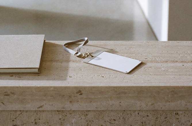 A blank visitor badge on a grey lanyard beside a closed linen-bound book on a travertine reception counter, an empty lobby behind.